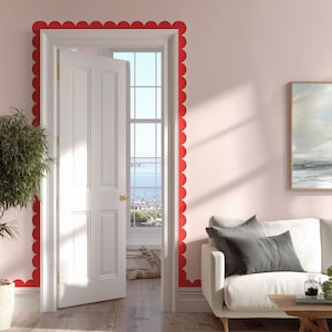 May include: A white door, slightly open, offers a view of the sea through a window. The door frame is adorned with a scalloped red border. The room features pale pink walls, a white sofa, and a framed picture.
