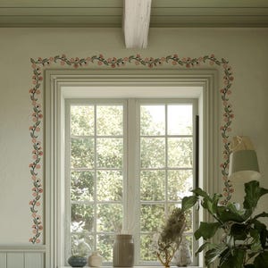 May include: A light green wall with a white window frame, adorned with a floral garland. The garland features pink flowers and green leaves. A wooden beam is visible above the window. The room is decorated with plants and a lamp.
