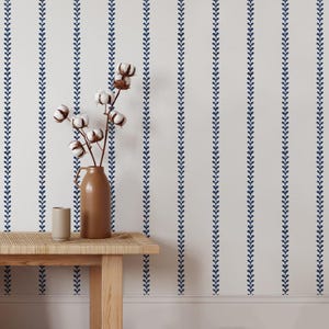 May include: A white wall with a repeating pattern of blue vertical lines of small, stylized leaves. A wooden table with a vase of white cotton flowers sits in front of the wall.