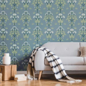 May include: A blue wallpaper with a repeating floral pattern in white and green. A gray sofa with a striped blanket and a decorative pillow is in front of the wallpaper. A white vase sits on a wooden side table.