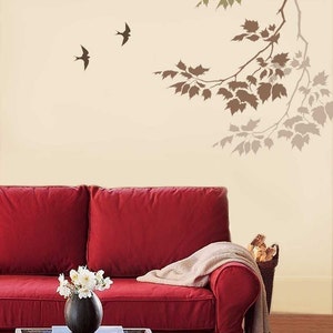 May include: A red sofa with a white throw blanket and a vase of white flowers on a coffee table. The wall behind the sofa has a stencil of a tree branch with leaves and two birds.