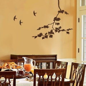 May include: A dining room with a brown wooden table and chairs. The wall is painted a light yellow and features a brown silhouette of a tree branch with leaves and birds.
