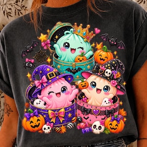 May include: A charcoal gray t-shirt featuring a graphic of three cute ghosts in colorful round boxes. One ghost wears a gold crown, while the others wear witch hats. The design includes orange pumpkins, small bats, skulls, stars, and pink bows.