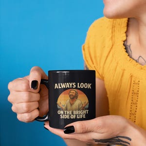 May include: Black ceramic mug featuring the text "ALWAYS LOOK ON THE BRIGHT SIDE OF LIFE" and an illustration of a smiling man. The mug has a black handle and is held by a person with black nail polish.