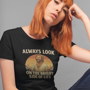 May include: Black t-shirt featuring the phrase "ALWAYS LOOK ON THE BRIGHT SIDE OF LIFE" and a graphic of a smiling man with outstretched arms against a sunset. The shirt is worn by a person with red hair.