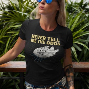 May include: Black t-shirt featuring the text "NEVER TELL ME THE ODDS" and a Millennium Falcon graphic. The shirt is worn with blue jeans and sunglasses.