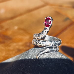 May include: A silver snake ring with a ruby-colored gemstone at the head. The ring is coiled and rests on a dark gray stone. The snake's body has a textured pattern. The background is a blurred wooden surface.