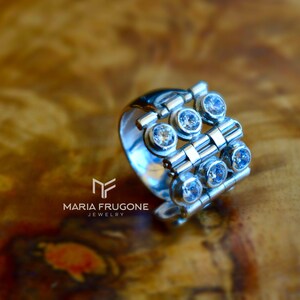 May include: A silver ring with a geometric design featuring multiple clear gemstones. The ring is made of linked metal pieces and has a modern, minimalist style.
