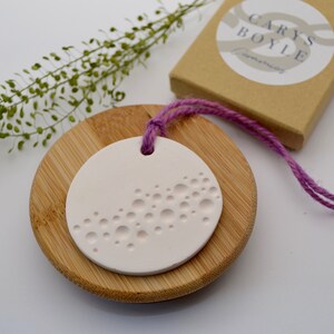 May include: A white ceramic ornament with a raised pattern of circles. The ornament is hanging from a pink string and is sitting on a wooden base.