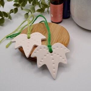 May include: Two off-white leaf-shaped ceramic ornaments with a dotted pattern and green twine hangers. The leaves rest on a wooden coaster, with essential oil bottles in the background. The ornaments are designed for aromatherapy.