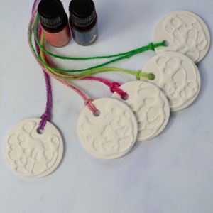 May include: Five white ceramic round ornaments with a raised design, each with a hole in the centre and attached to a colourful string. The strings are pink, green, and purple. Two small glass bottles with labels are in the background.