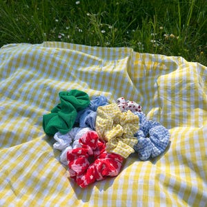 Pode incluir: Uma coleção de scrunchies coloridos em vários padrões e cores, incluindo verde, vermelho, amarelo e azul. Os scrunchies estão dispostos sobre uma manta de piquenique xadrez amarela e branca, com um fundo gramado.
