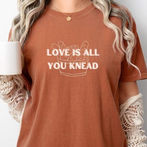 May include: A rust-coloured t-shirt with the text "LOVE IS ALL YOU KNEAD" in white. The shirt is paired with blue jeans. A white mug is held in the left hand. The overall style is casual and comfortable.