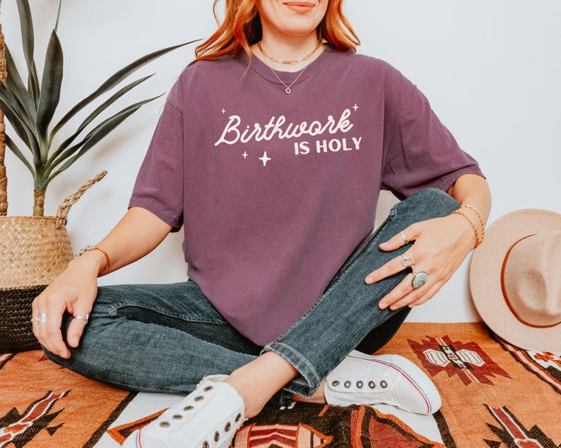 May include: A woman wearing a purple t-shirt with the text "Birthwork is Holy" in white lettering. She is wearing blue jeans and white sneakers.