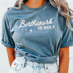 May include: A blue t-shirt with the text "Birthwork is Holy" in white lettering. The shirt is tied at the waist and the model is wearing light blue jeans.