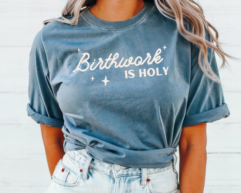 May include: A blue t-shirt with the text "Birthwork is Holy" in white lettering. The shirt is tied at the waist and the model is wearing light blue jeans.