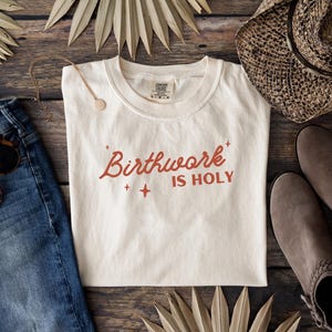 May include: A white t-shirt with the text "Birthwork is Holy" in red lettering. The shirt is laid flat on a wooden surface with a pair of jeans, a hat, and palm leaves.