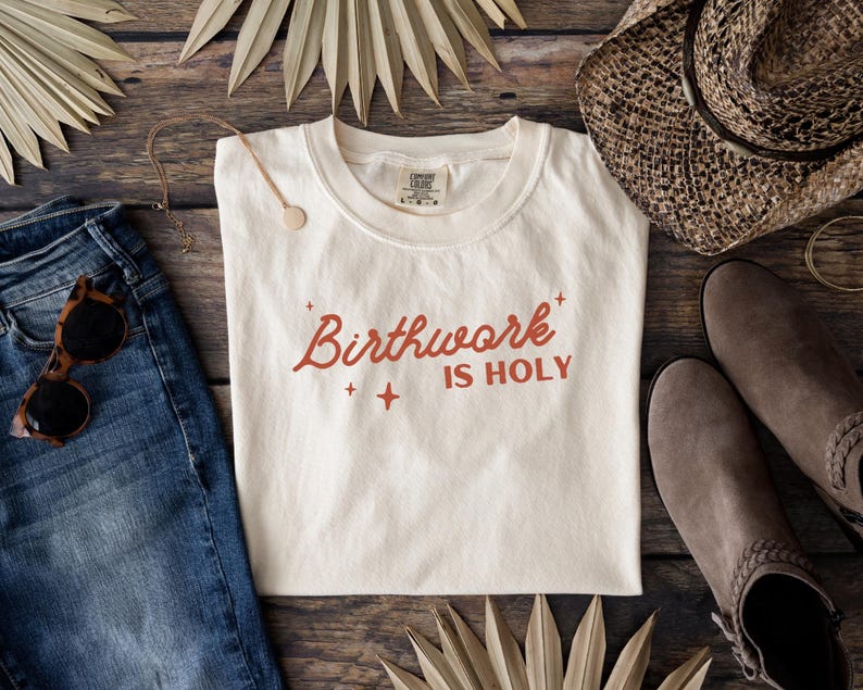 May include: A white t-shirt with the text "Birthwork is Holy" in red lettering. The shirt is laid flat on a wooden surface with a pair of jeans, a hat, and palm leaves.