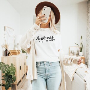 May include: A white t-shirt with the text "Birthwork is Holy" in black lettering. The shirt is being worn by a person with a brown hat and a white cardigan.