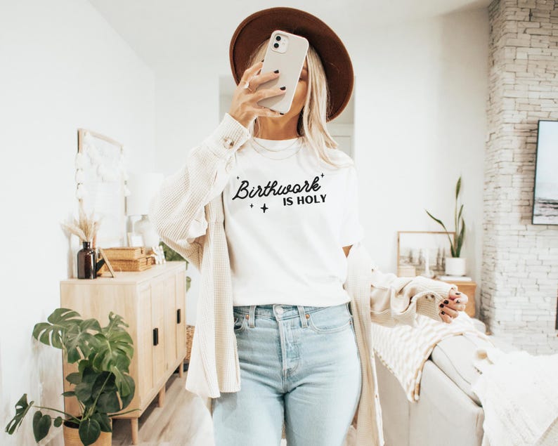 May include: A white t-shirt with the text "Birthwork is Holy" in black lettering. The shirt is being worn by a person with a brown hat and a white cardigan.
