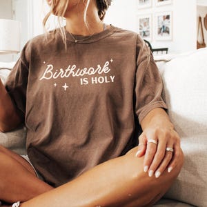 May include: A brown t-shirt with the text "Birthwork is Holy" in white lettering. The shirt is worn by a person sitting on a couch.