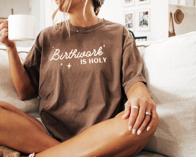 May include: A brown t-shirt with the text "Birthwork is Holy" in white lettering. The shirt is worn by a person sitting on a couch.