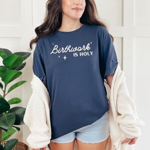 May include: A navy blue t-shirt with the text "Birthwork is Holy" in white lettering. The shirt has a relaxed fit and is being worn by a person with long brown hair.
