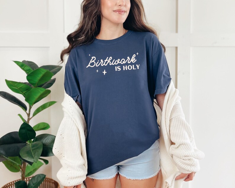 May include: A navy blue t-shirt with the text "Birthwork is Holy" in white lettering. The shirt has a relaxed fit and is being worn by a person with long brown hair.