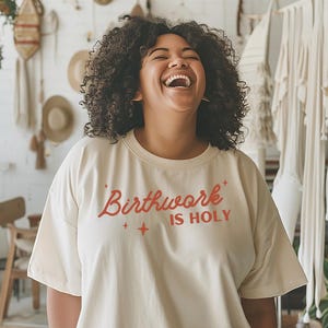 May include: A woman wearing a cream colored t-shirt with the text "Birthwork is Holy" in orange lettering. She is laughing and has curly dark hair.