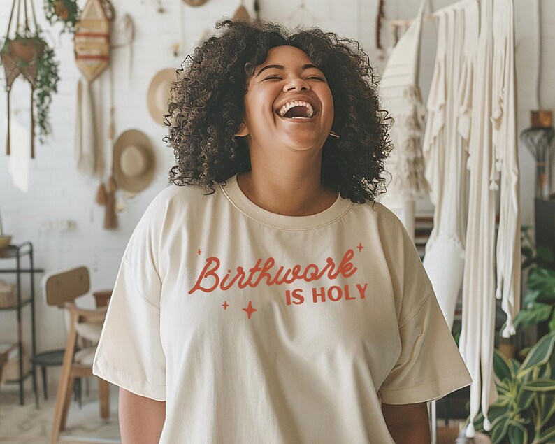 May include: A woman wearing a cream colored t-shirt with the text "Birthwork is Holy" in orange lettering. She is laughing and has curly dark hair.