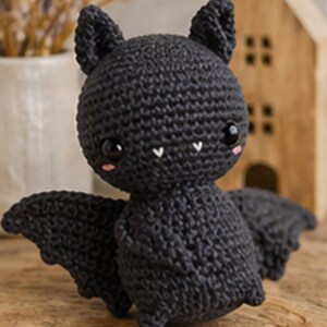 Peut inclure: Une chauve-souris noire au crochet faite &agrave; la main, avec de grands yeux expressifs, de petites joues roses et des crocs blancs. La chauve-souris a des ailes d&eacute;ploy&eacute;es et est pos&eacute;e sur une surface en bois. Une petite maison en bois est en arri&egrave;re-plan.