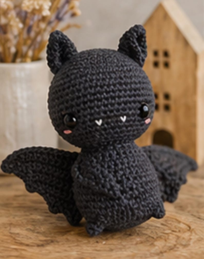 Peut inclure: Une chauve-souris noire au crochet faite &agrave; la main, avec de grands yeux expressifs, de petites joues roses et des crocs blancs. La chauve-souris a des ailes d&eacute;ploy&eacute;es et est pos&eacute;e sur une surface en bois. Une petite maison en bois est en arri&egrave;re-plan.