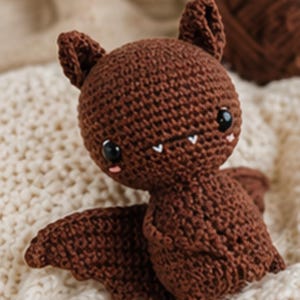 Peut inclure: Une poup&eacute;e chauve-souris au crochet marron faite &agrave; la main, avec de grands yeux noirs, de petites joues roses et des crocs blancs. La chauve-souris a de grandes oreilles et des ailes d&eacute;ploy&eacute;es. La poup&eacute;e est pos&eacute;e sur une surface textur&eacute;e de couleur cr&egrave;me.