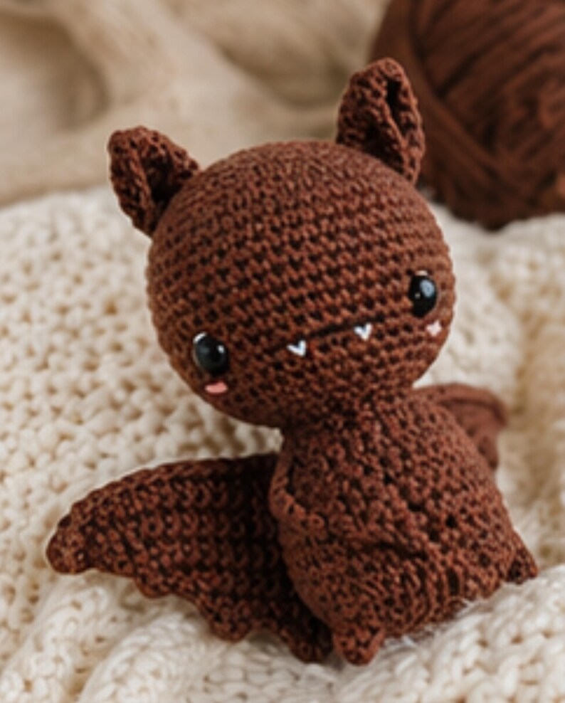 Peut inclure: Une poup&eacute;e chauve-souris au crochet marron faite &agrave; la main, avec de grands yeux noirs, de petites joues roses et des crocs blancs. La chauve-souris a de grandes oreilles et des ailes d&eacute;ploy&eacute;es. La poup&eacute;e est pos&eacute;e sur une surface textur&eacute;e de couleur cr&egrave;me.