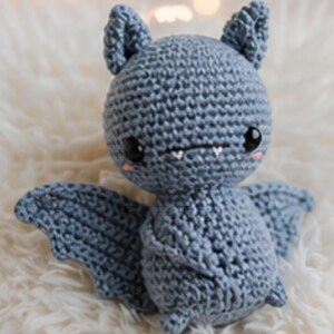Peut inclure: Une peluche chauve-souris au crochet bleu clair. Le jouet fait main pr&eacute;sente de grands yeux noirs, de petites dents et des joues roses. La chauve-souris a des ailes et des oreilles d&eacute;taill&eacute;es, pos&eacute;e sur une surface blanche et moelleuse.