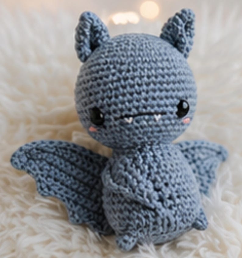 Peut inclure: Une peluche chauve-souris au crochet bleu clair. Le jouet fait main pr&eacute;sente de grands yeux noirs, de petites dents et des joues roses. La chauve-souris a des ailes et des oreilles d&eacute;taill&eacute;es, pos&eacute;e sur une surface blanche et moelleuse.
