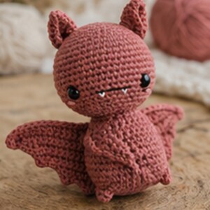 Peut inclure: Une chauve-souris au crochet de couleur rose poudr&eacute;. La chauve-souris a de grands yeux noirs, de petites dents blanches et des ailes d&eacute;ploy&eacute;es. La chauve-souris est pos&eacute;e sur une surface en bois.