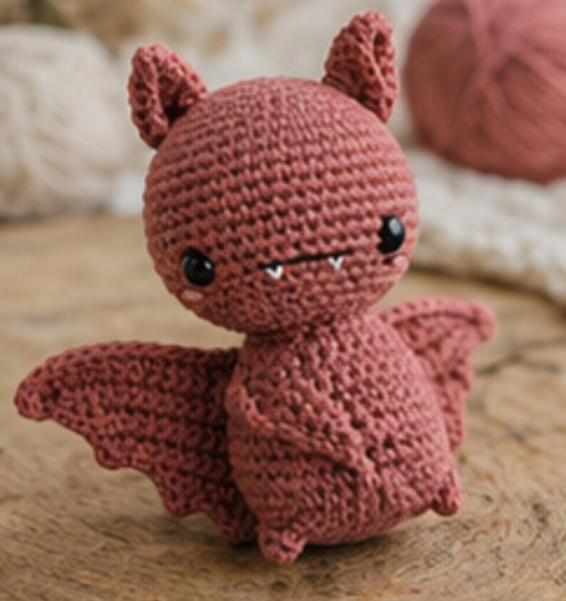 Peut inclure: Une chauve-souris au crochet de couleur rose poudr&eacute;. La chauve-souris a de grands yeux noirs, de petites dents blanches et des ailes d&eacute;ploy&eacute;es. La chauve-souris est pos&eacute;e sur une surface en bois.