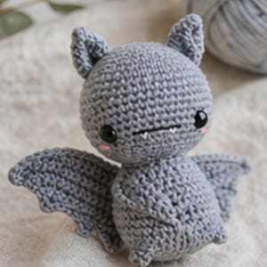 Peut inclure: Une chauve-souris grise au crochet faite &agrave; la main, avec de grands yeux noirs, de petites canines et des joues roses. La chauve-souris a des ailes d&eacute;ploy&eacute;es et de petites oreilles. Une pelote de laine grise est en arri&egrave;re-plan. C'est un jouet mignon et fantaisiste.