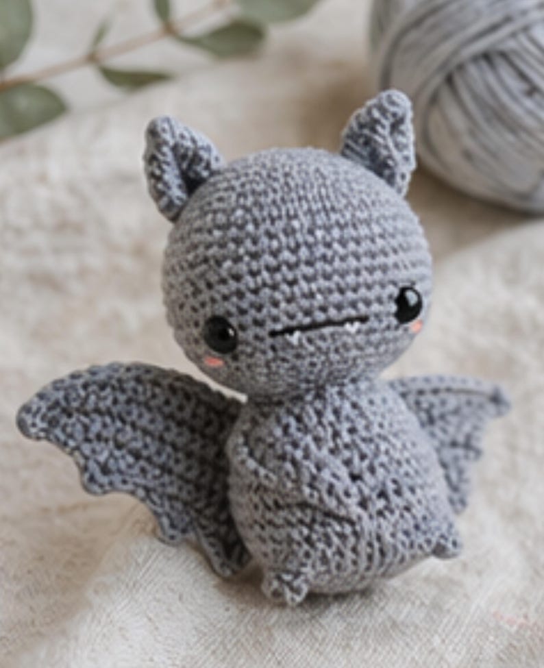 Peut inclure: Une chauve-souris grise au crochet faite &agrave; la main, avec de grands yeux noirs, de petites canines et des joues roses. La chauve-souris a des ailes d&eacute;ploy&eacute;es et de petites oreilles. Une pelote de laine grise est en arri&egrave;re-plan. C'est un jouet mignon et fantaisiste.