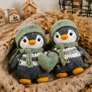Peut inclure: Deux poupées pingouins faites à la main au crochet. Chaque pingouin porte un chapeau et une écharpe verts, avec un corps rayé blanc et gris. Un pingouin tient un cœur vert. Les pingouins ont des becs et des pieds orange. Le fond comprend de petites maisons en bois et une couverture tissée.