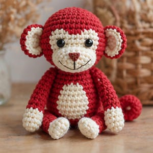 Könnte beinhalten: Handgemachte Häkelpuppe eines Affen in Rot und Creme. Der Affe hat ein lächelndes Gesicht, schwarze Augen und einen cremefarbenen Bauch und Füße. Die Ohren und Hände sind ebenfalls cremefarben. Eine kleine rote Kugel liegt neben dem Affen.