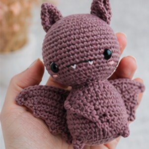 Peut inclure: Une chauve-souris crochet&eacute;e de couleur rose poussi&eacute;reux. La chauve-souris a de grands yeux noirs, de petites dents blanches et des oreilles pointues. Ses ailes sont d&eacute;ploy&eacute;es, et elle est tenue dans une main. Le jouet fait main est un design mignon et fantaisiste.