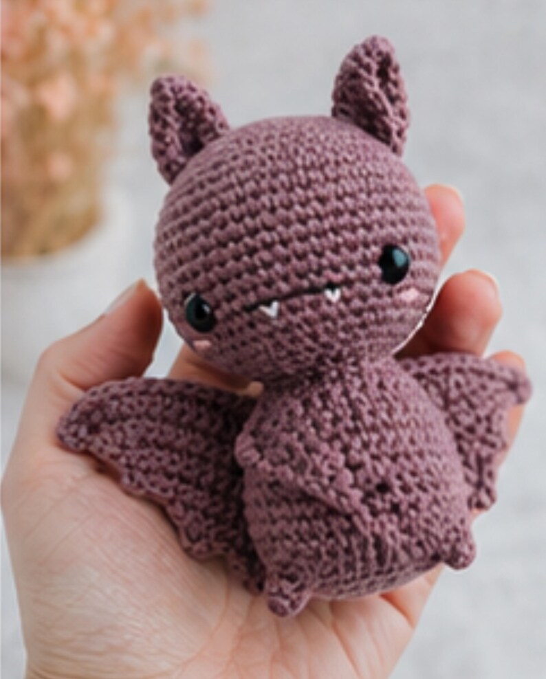 Peut inclure: Une chauve-souris crochet&eacute;e de couleur rose poussi&eacute;reux. La chauve-souris a de grands yeux noirs, de petites dents blanches et des oreilles pointues. Ses ailes sont d&eacute;ploy&eacute;es, et elle est tenue dans une main. Le jouet fait main est un design mignon et fantaisiste.