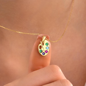 May include: A gold necklace with a pendant in the shape of an artist's palette. The palette has gemstones in various colours. The necklace is on a fine gold chain, making it a stylish and artistic piece of jewellery.