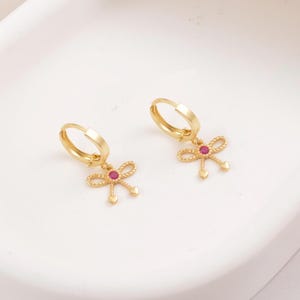 May include: A pair of gold-colored hoop earrings. Each earring features a bow-shaped charm with a small red gem in the center and two heart-shaped dangles. The earrings are displayed on a white surface.