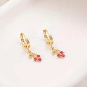 May include: A pair of gold-colored hoop earrings. Each earring features a dangling charm with three red gemstones, a green gemstone, and a small gold leaf. The earrings are displayed on a white surface.