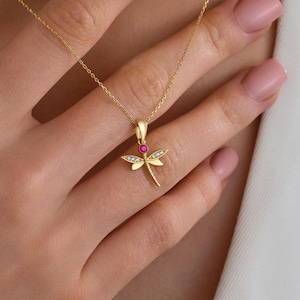 May include: A gold-colored dragonfly pendant necklace. The dragonfly has a red gemstone body and diamond-like accents on the wings. The necklace is on a delicate gold chain.