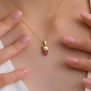 May include: A gold cat pendant necklace featuring a red heart-shaped gemstone. The necklace is suspended from a fine gold chain. The cat pendant has a small loop at the top for the chain to pass through.