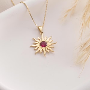 May include: Gold-toned necklace featuring a sun pendant with a central, round, red gemstone. The sunburst design has pointed rays. The necklace is displayed on a white surface, with a soft, neutral background.